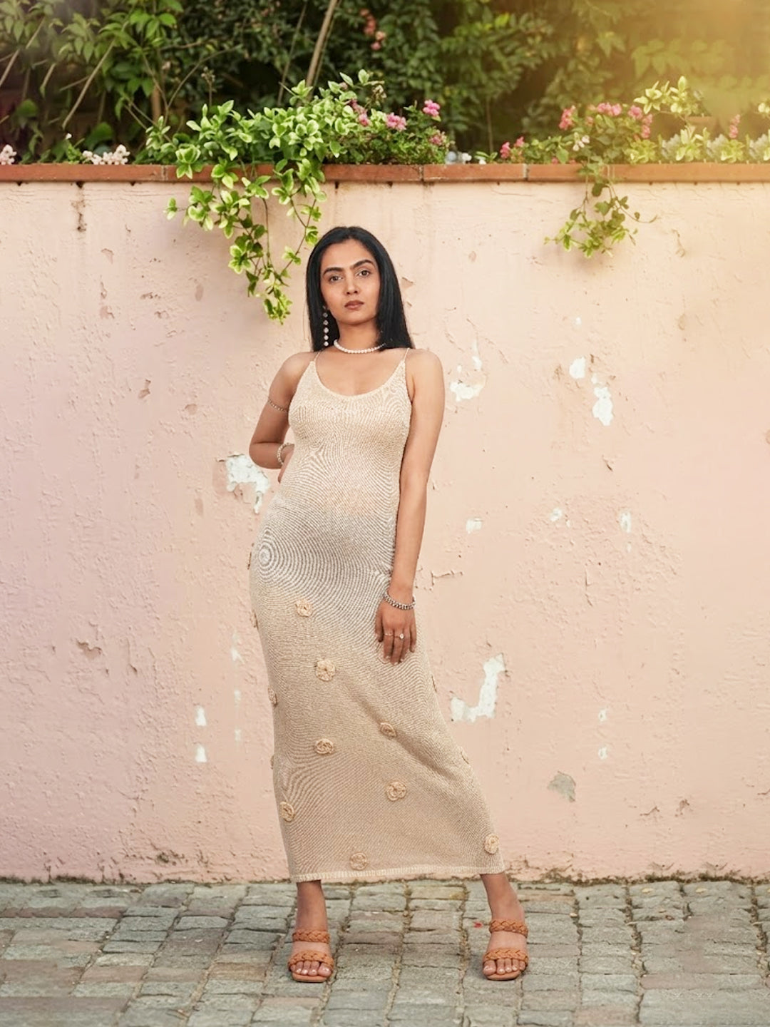 Woman in a beige dress standing against a light pink wall with greenery.