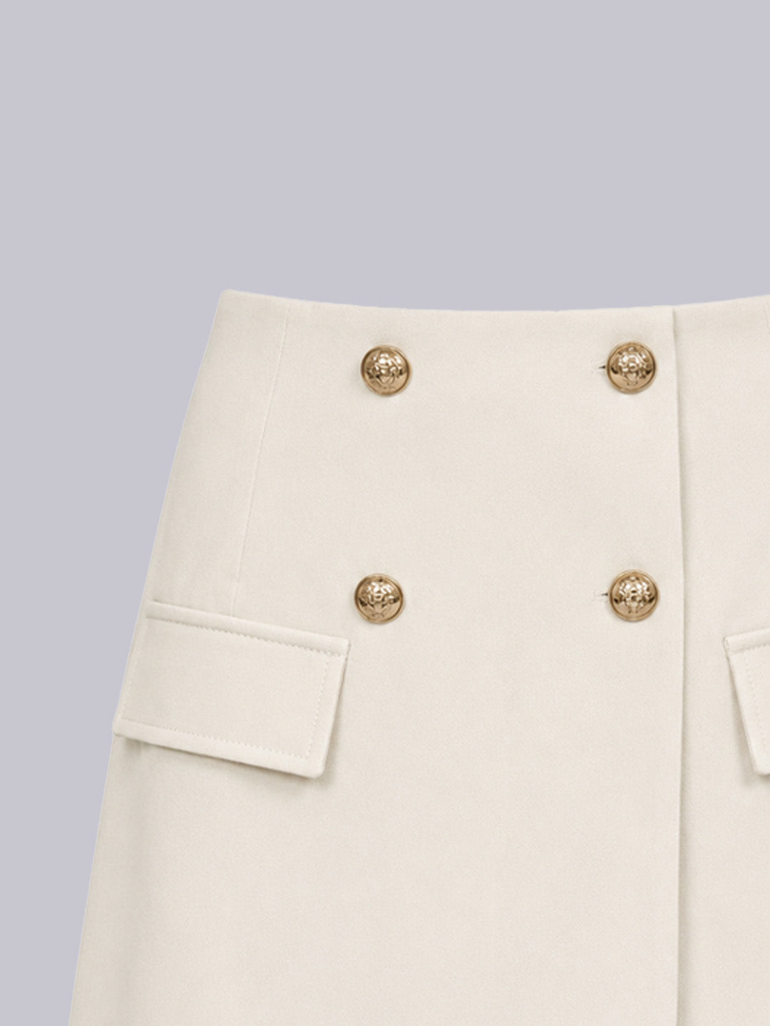 Close-up of a cream-colored skirt featuring a double-breasted design with four gold-tone decorative buttons and flap pockets on the sides.