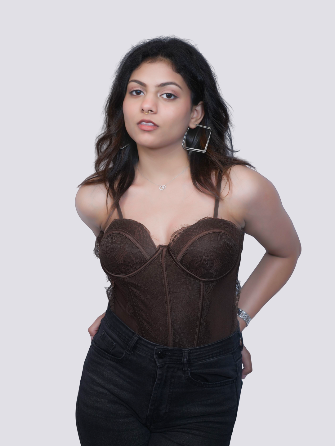 A woman modeling a dark brown lace and mesh corset-style camisole top with underwire cups, paired with black jeans, against a solid light gray background.