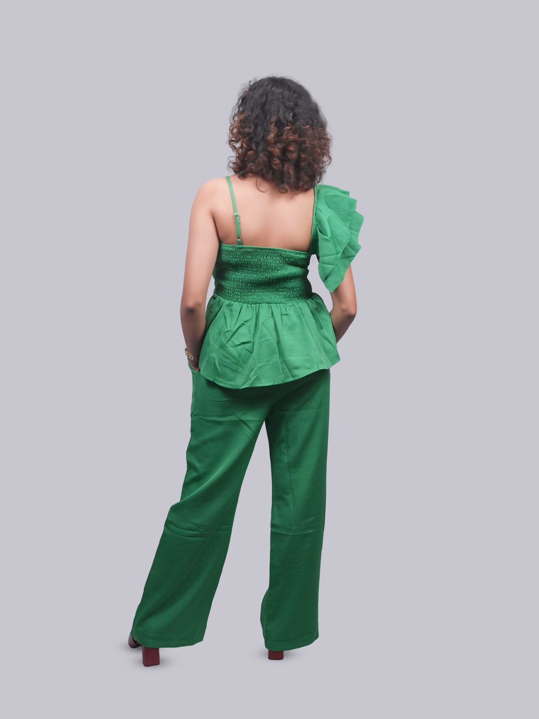 A model shown from the back wearing a green two-piece outfit consisting of a one-shoulder smocked peplum top and matching wide-leg pants, set against a solid gray background.