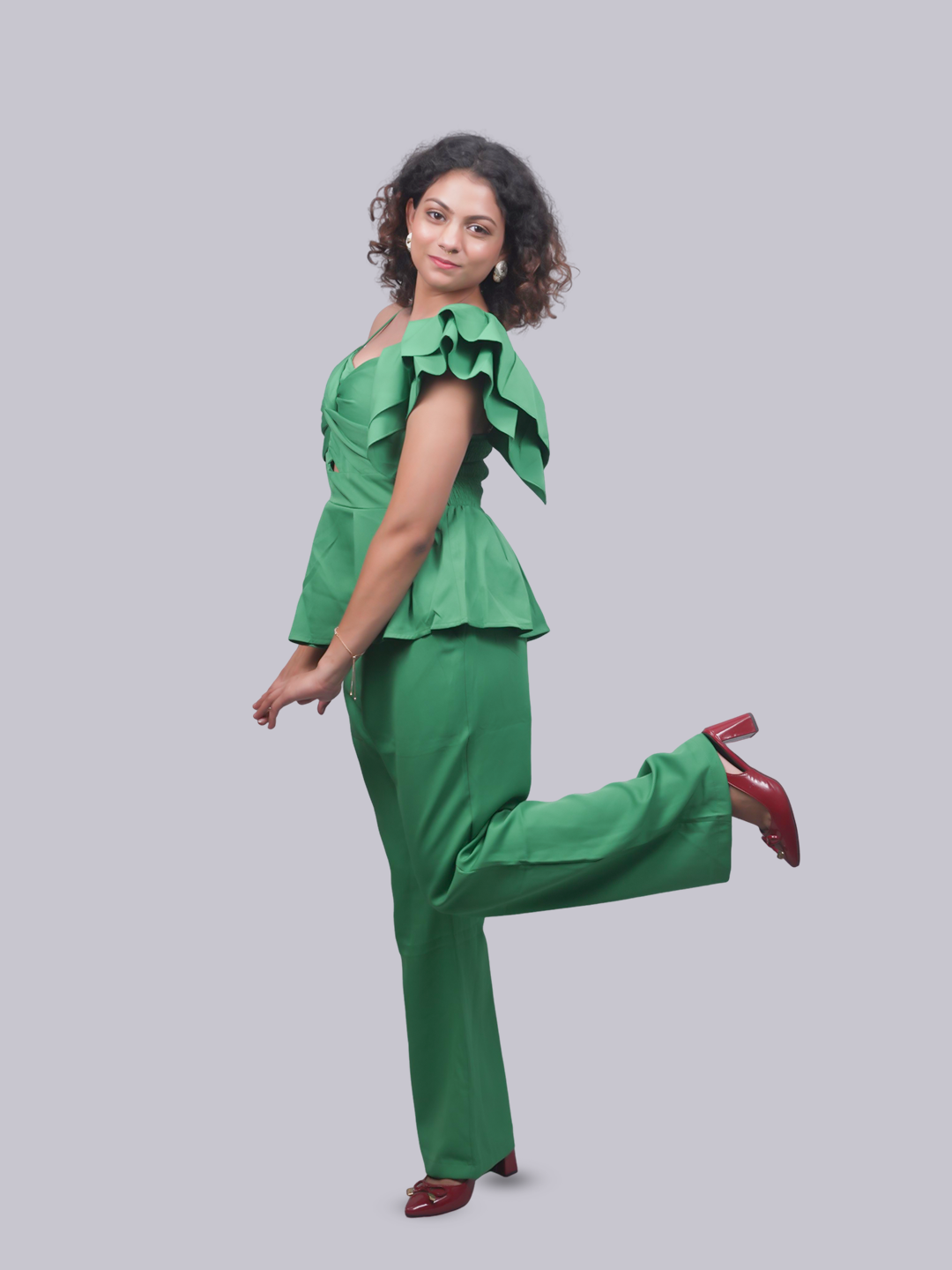 A woman modeling a bright green two-piece outfit consisting of a ruffled off-the-shoulder peplum top and matching wide-leg pants, paired with dark red heels, against a solid gray background.