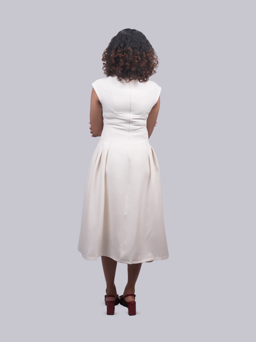 A woman viewed from behind wearing a sleeveless cream-colored midi dress with a gathered waist and pleated skirt, paired with dark brown block-heel sandals, against a solid light gray background.