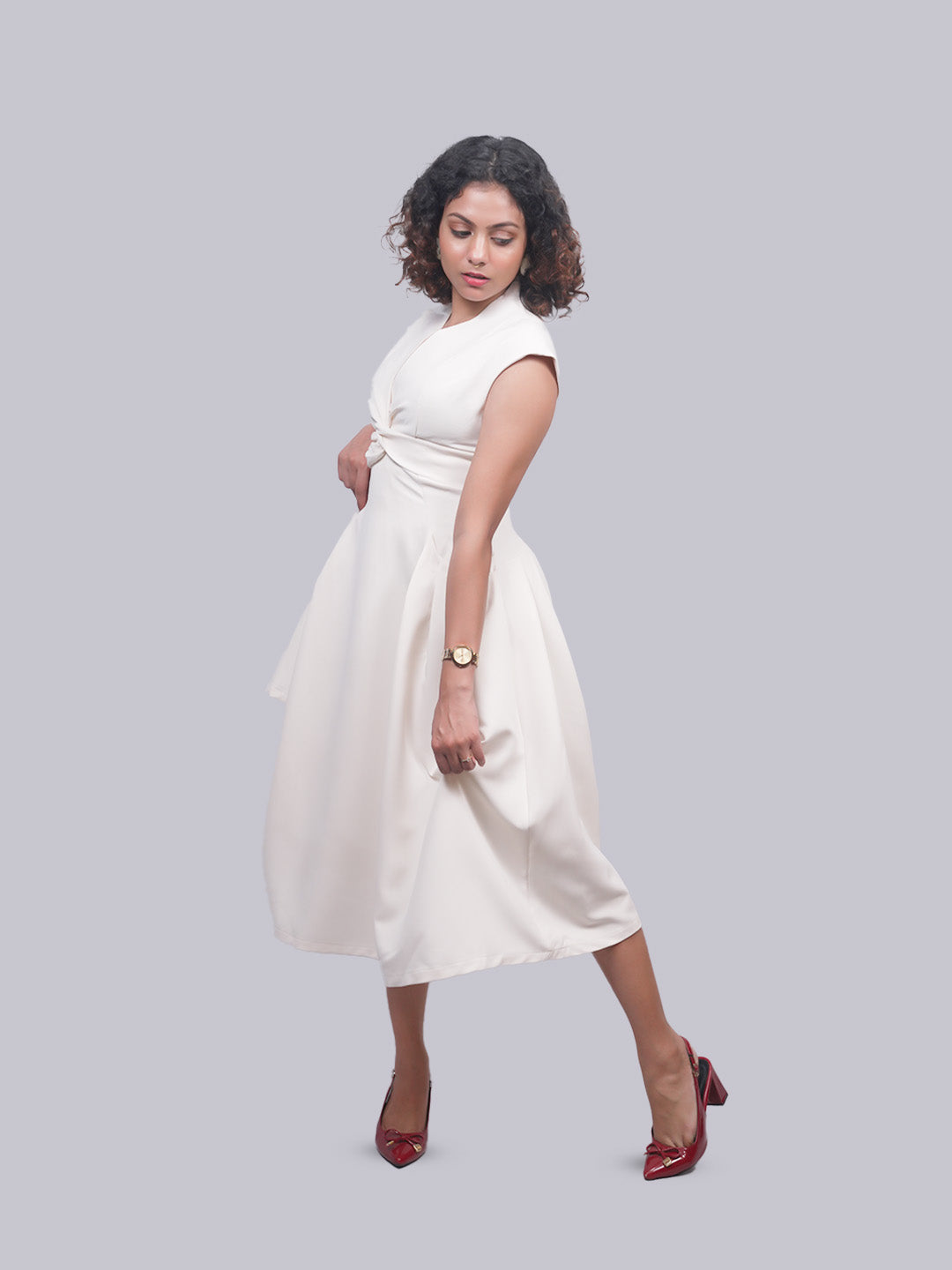 A woman modeling a sleeveless white midi dress with a wrap-style bodice and flared skirt, paired with dark red pointed heels, against a solid light gray background.