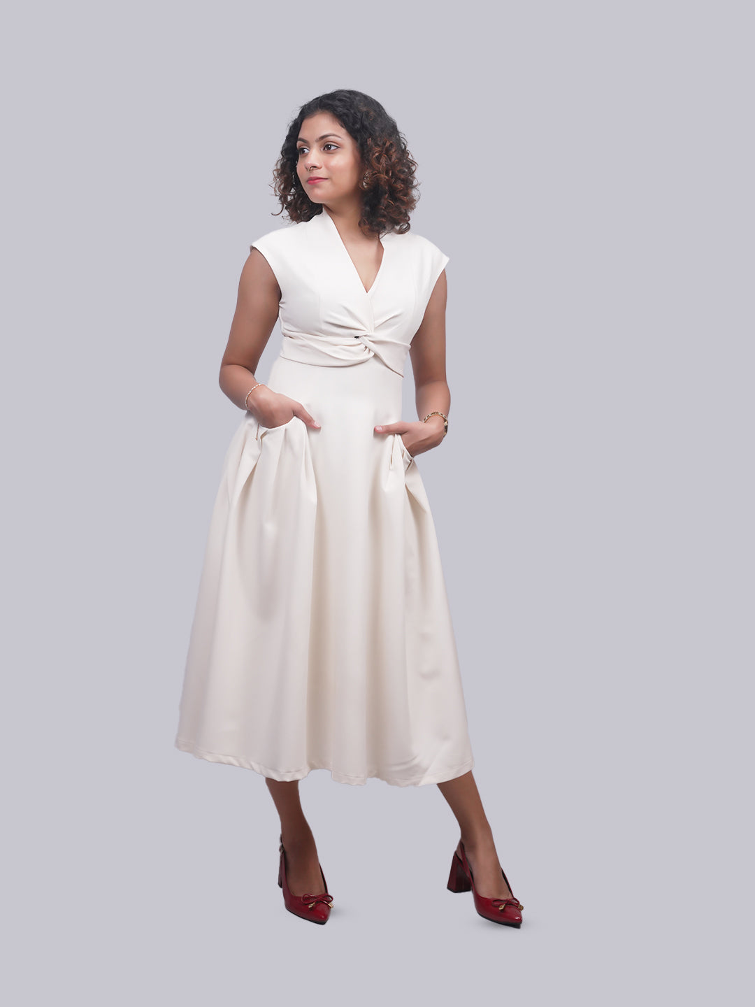 A woman modeling a sleeveless cream midi dress with a twisted waist detail and pockets, paired with dark red pointed-toe heels, against a solid gray background.