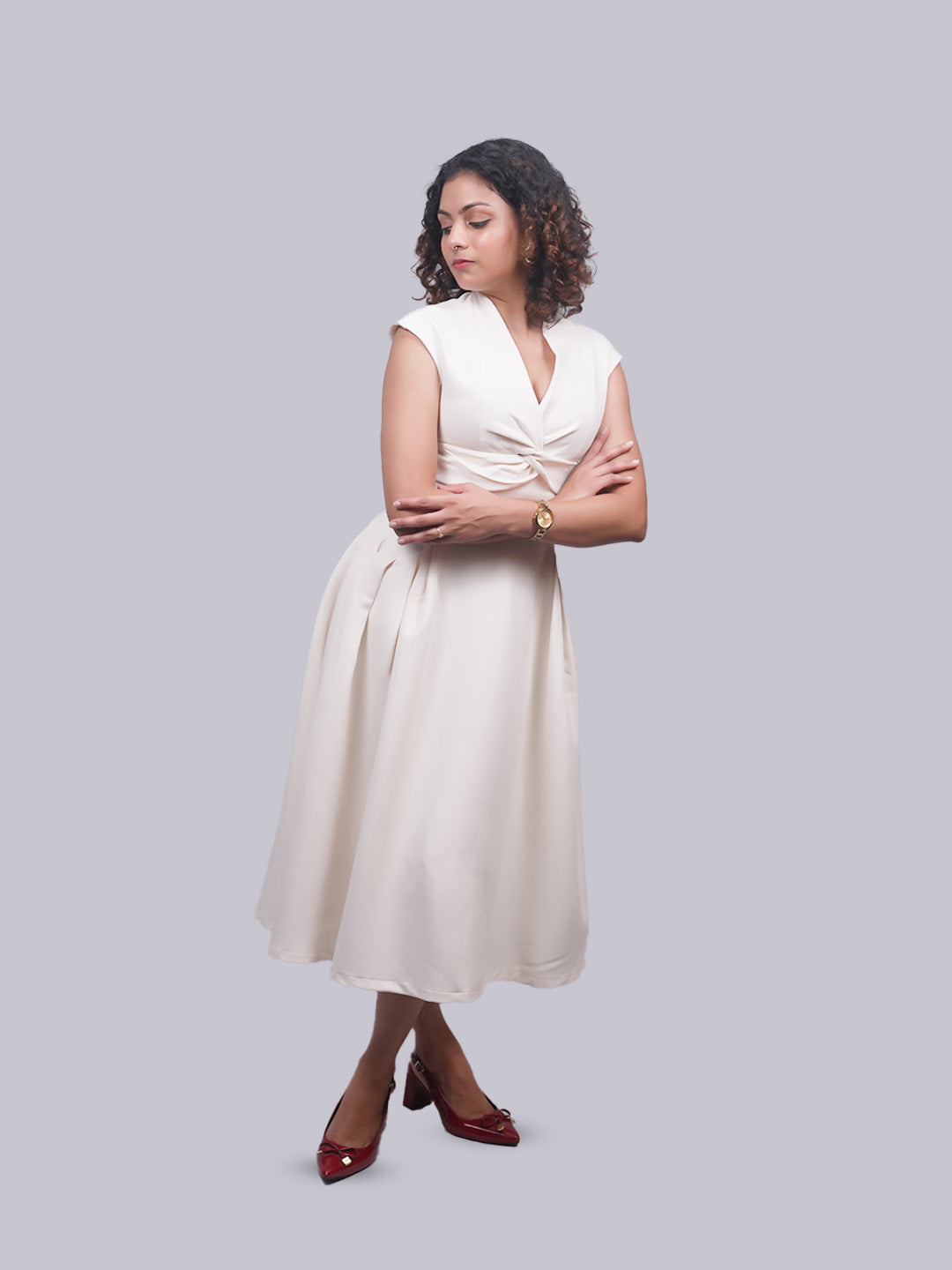 A woman modeling a sleeveless cream-colored midi dress with a twisted V-neck bodice, standing against a solid light gray background.
