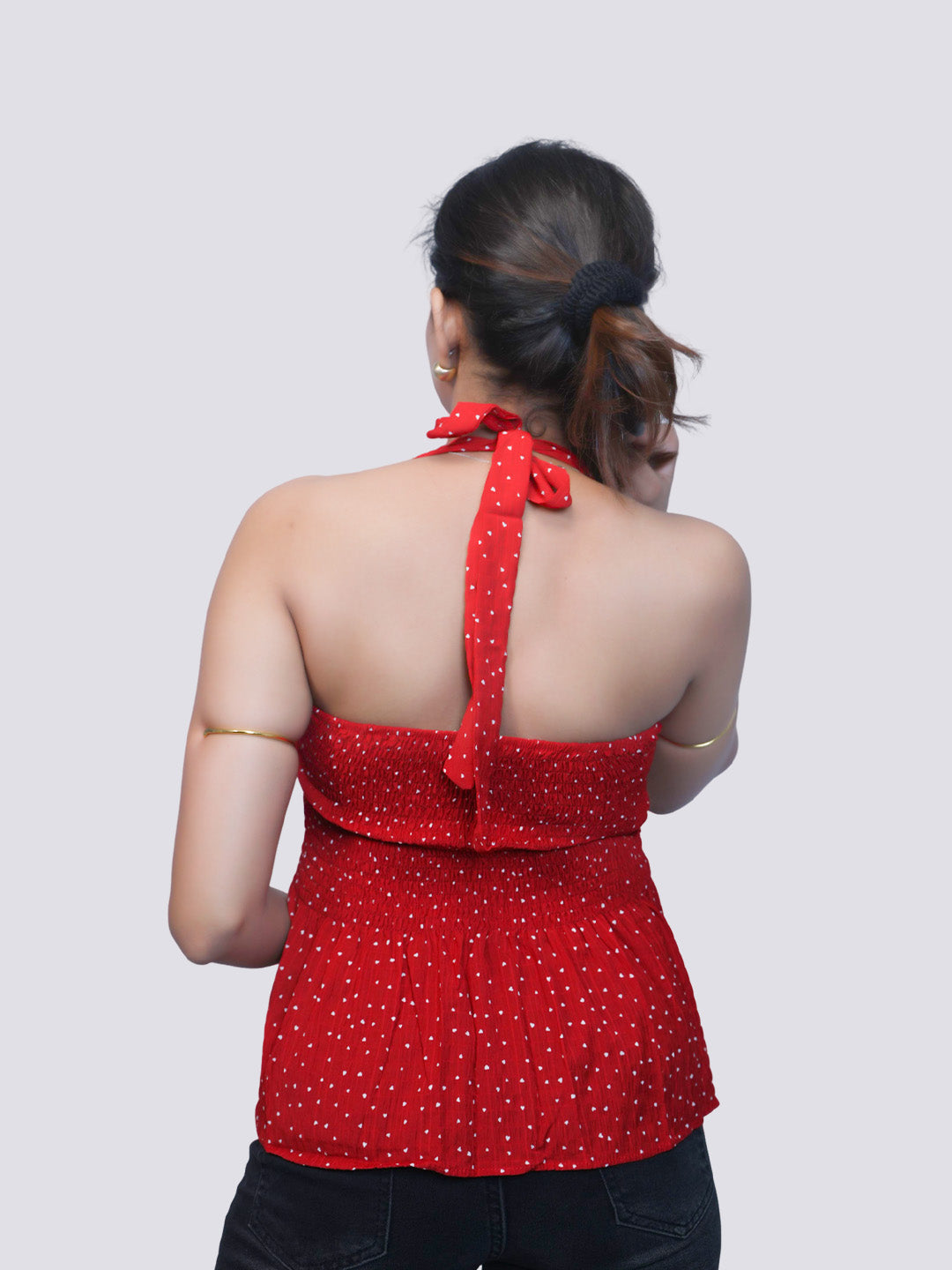 A woman shown from the back wearing a red halter-style top with a small white polka dot pattern and a tie-neck detail, paired with black pants, against a light gray studio background.