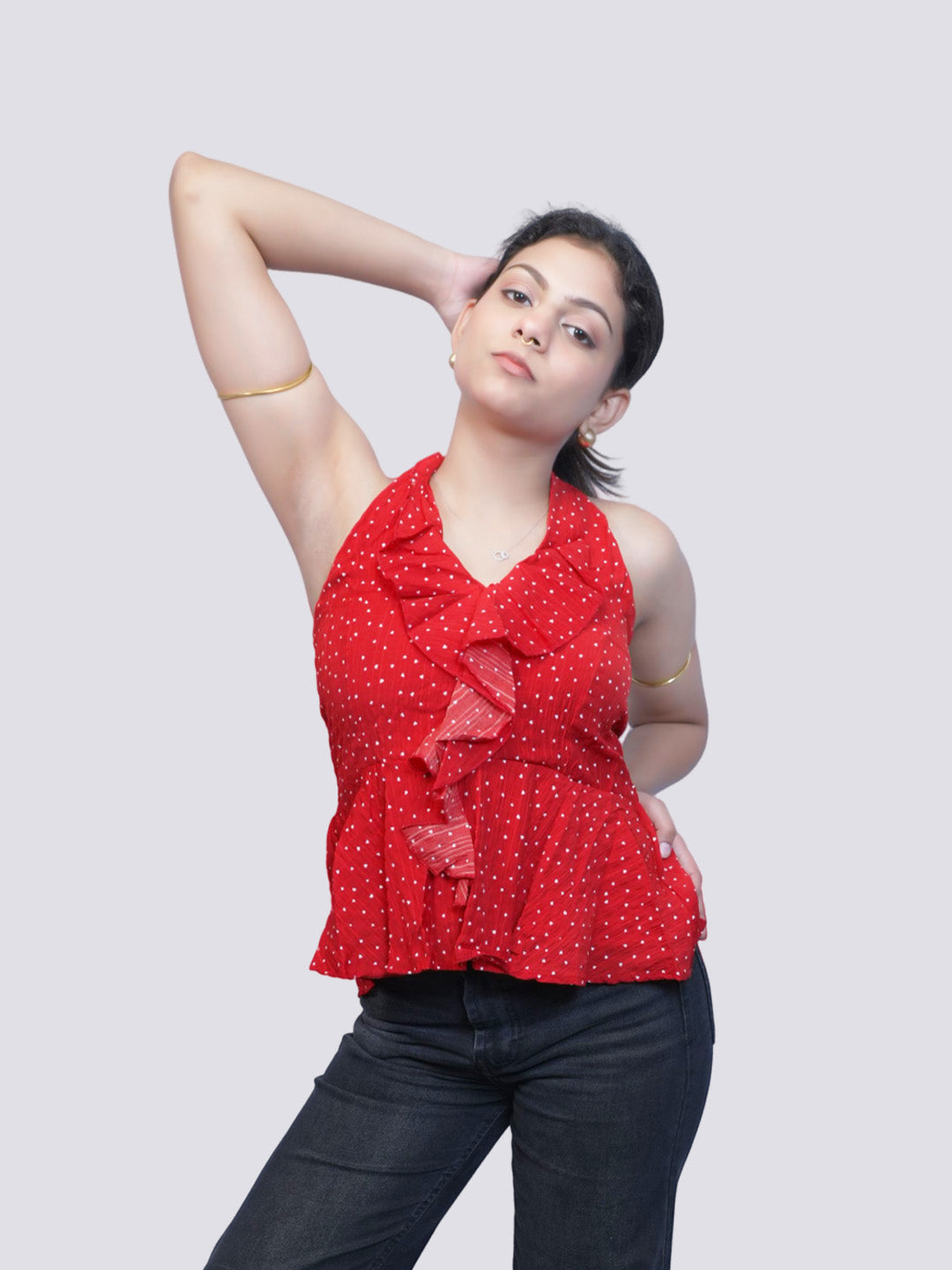 A woman modeling a red sleeveless ruffled top with a small white polka dot pattern, paired with dark jeans, against a solid light gray background.