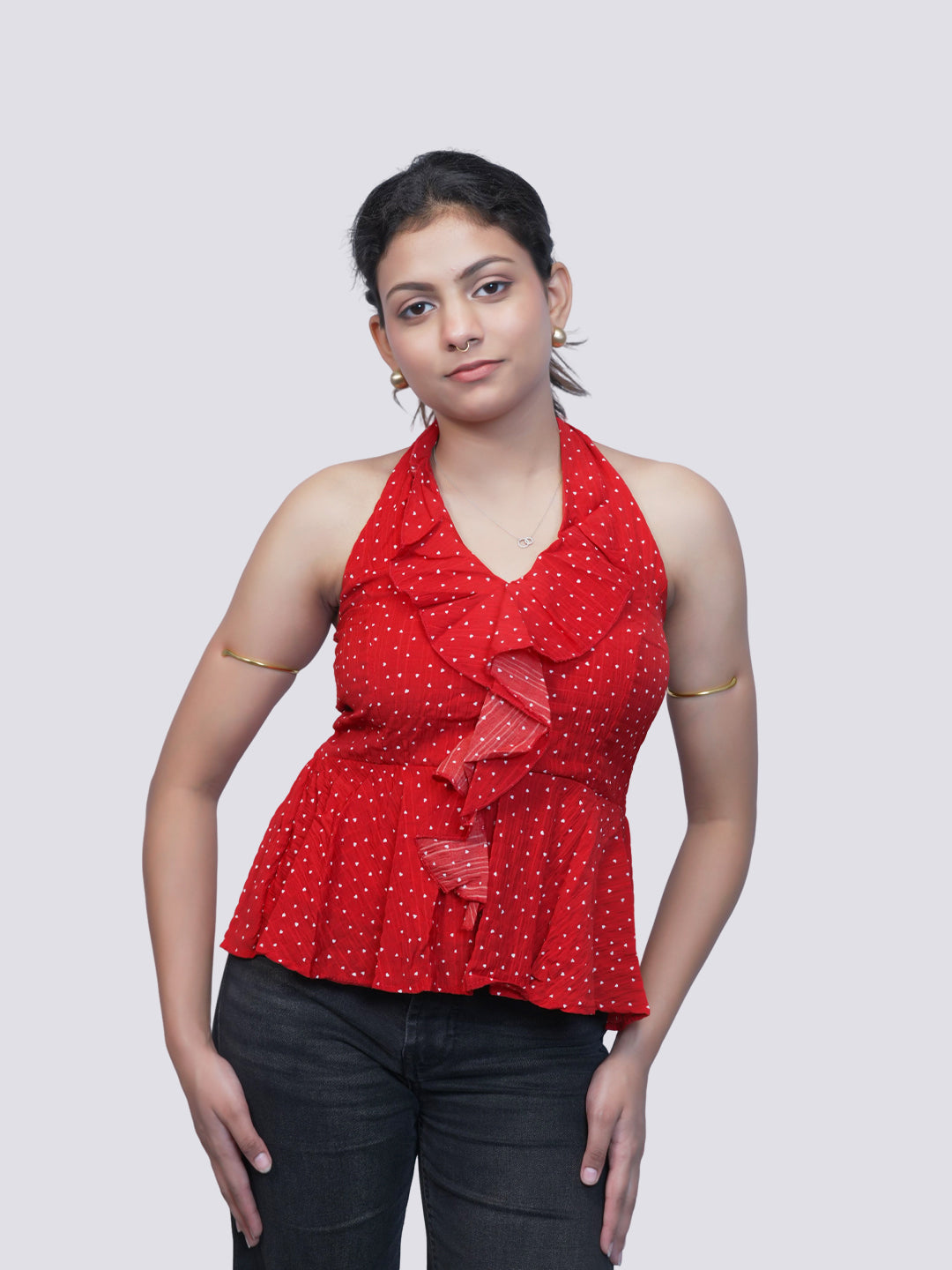 A woman modeling a red halter-neck top with white polka dots and ruffle details, paired with dark jeans, against a solid light gray background.
