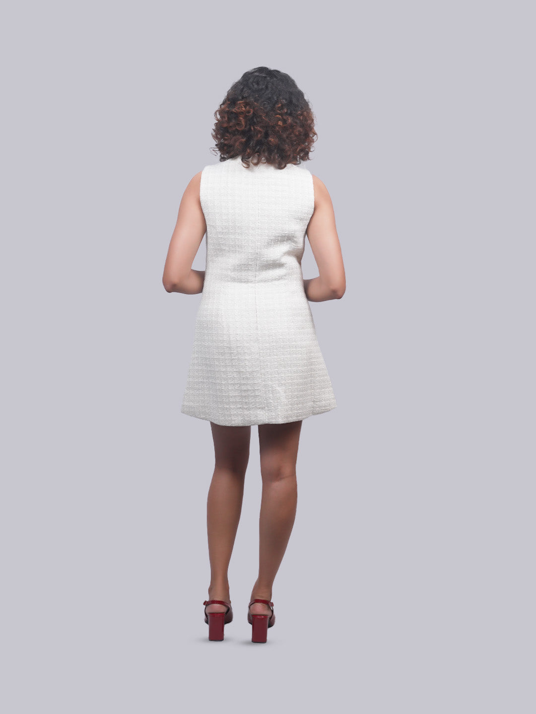 A woman viewed from behind wearing a sleeveless white textured mini dress and dark brown high heels, set against a solid light gray background.